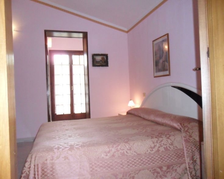 Einfaches Zimmer mit rosa Wänden, einem Doppelbett, sanfter Beleuchtung und Blick auf einen Garten.