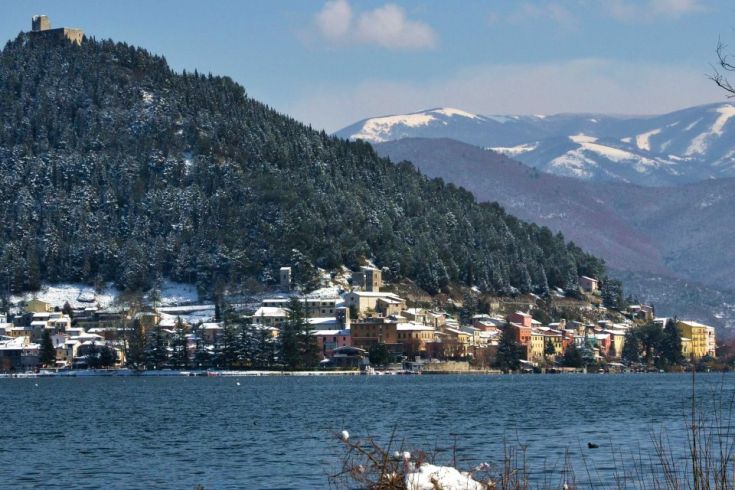 Winterpanorama mit einem See im Vordergrund und einem ruhigen Dorf im Hintergrund.