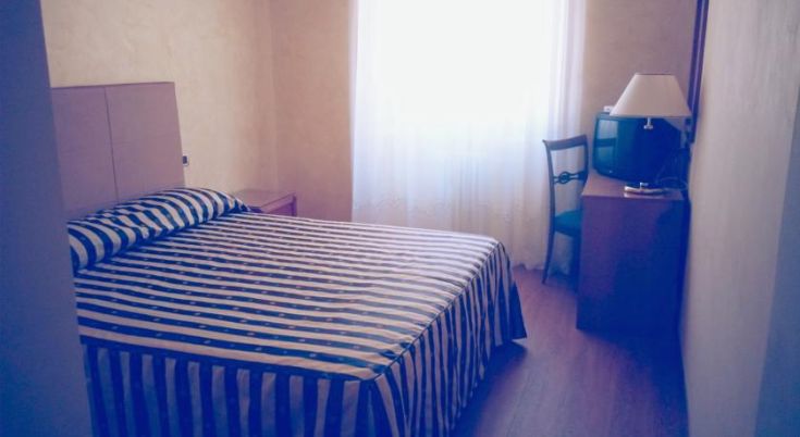 Camera semplice con letto a righe, TV e luce naturale che entra da una finestra ampia.