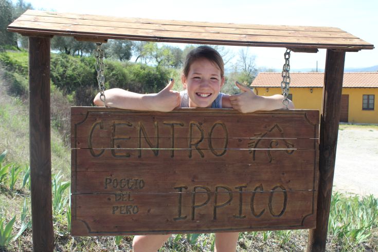 Una ragazza sorridente si trova davanti al cartello del centro ippico, circondato da un ambiente naturale.