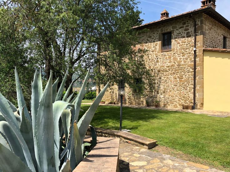 Casa colonica in pietra circondata da un giardino e da un paesaggio collinare.