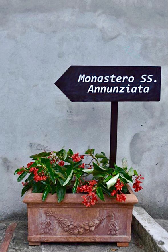 Ein Schild zeigt das Kloster SS. Annunziata, umgeben von Pflanzen mit roten Blumen.