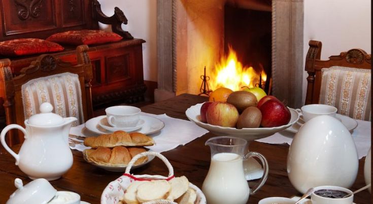 Colazione in una cucina confortevole, con camino acceso, frutta fresca e dolci sul tavolo.