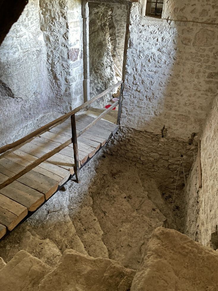 Innenraum einer antiken Struktur mit Steintreppen und einer Holzbrücke, die zu einem größeren Bereich führt.