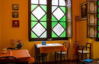 Interno di un ristorante con pareti gialle, finestre decorate e tavoli allestiti per i clienti, in un ambiente conviviale.