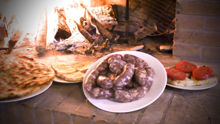 Piatti tipici serviti vicino al caminetto, con salsicce, pane e pomodori. Ambientazione accogliente e rustica.