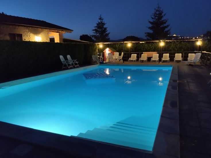 Piscina illuminata di notte, con lettini per il relax e piante ben curate nei dintorni.