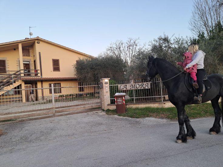 Una madre e sua figlia cavalcano un cavallo davanti a un agriturismo, circondate da un ambiente naturale.