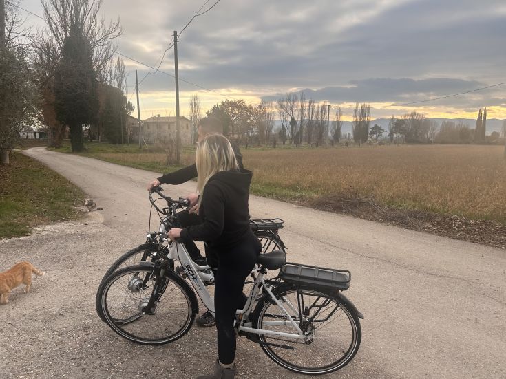 Due persone pedalano su biciclette elettriche lungo una strada di campagna verde, circondate dalla natura. Le nuvole coprono il cielo.