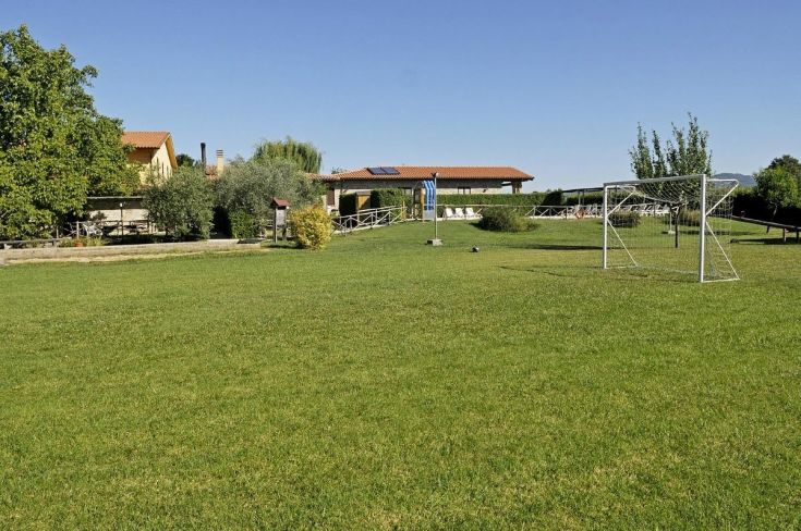 Un prato verde con un campo da calcetto, circondato da alberi e un edificio rustico sullo sfondo.