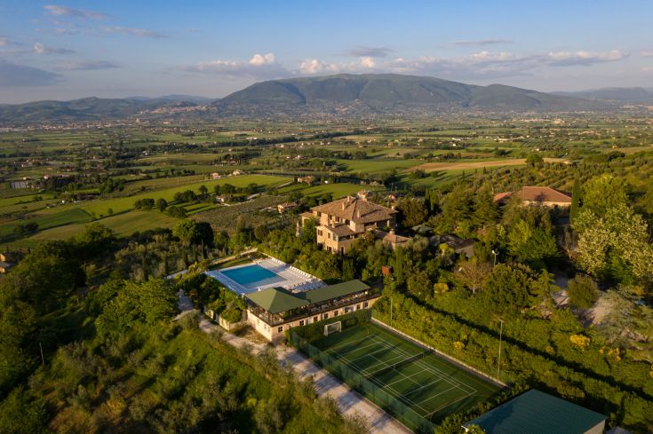 Scene of Poggio degli Olivi, surrounded by trees and featuring a pool, with sports fields visible in the background.