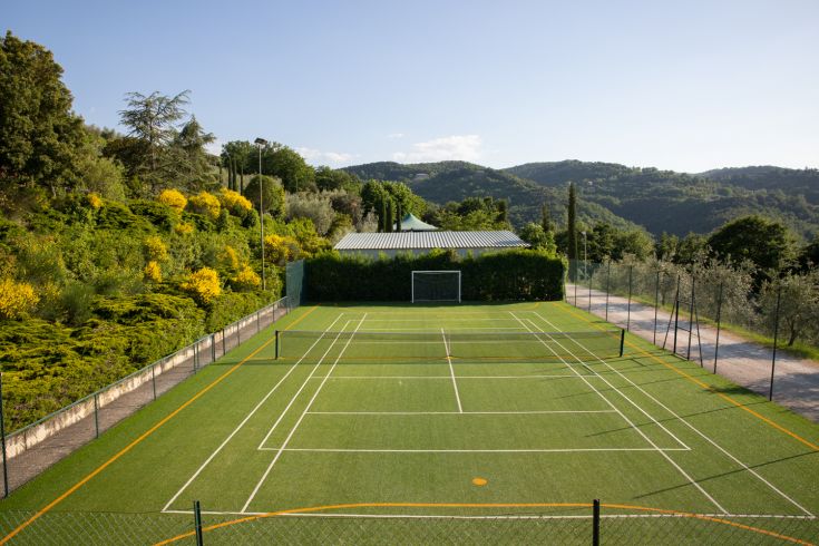 Tennis court located in a green area, surrounded by trees and hills.