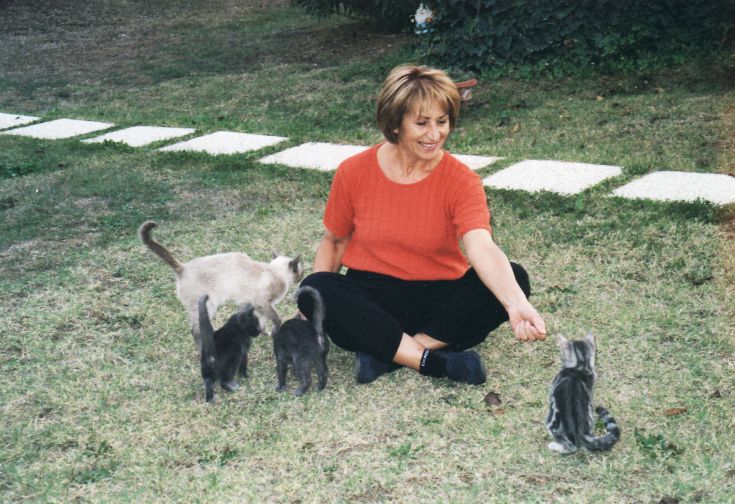 Eine Frau lächelt, während sie mit vier Katzen in einem hellgrünen Garten spielt.