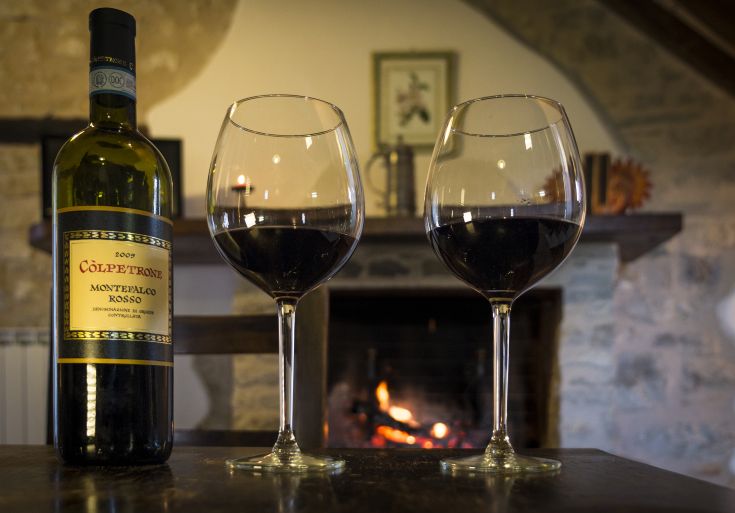 Una bottiglia di vino rosso di Montefalco e due bicchieri, con un camino acceso in secondo piano.