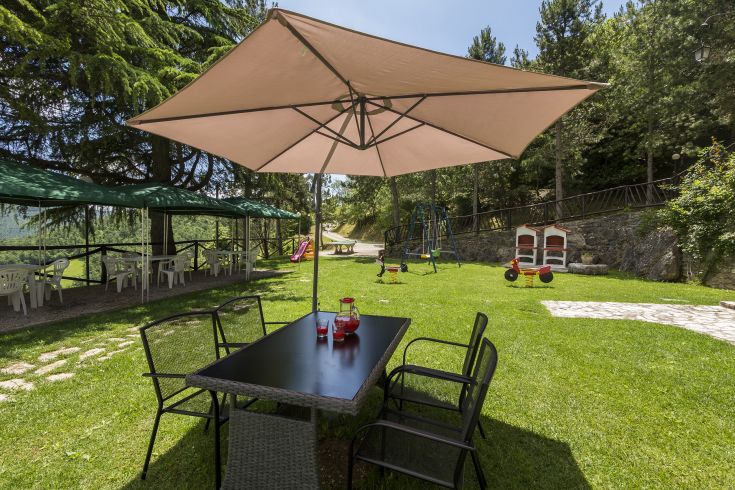 Giardino all'aperto, dotato di un tavolo, un ombrellone e giochi per bambini, circondato da vegetazione.