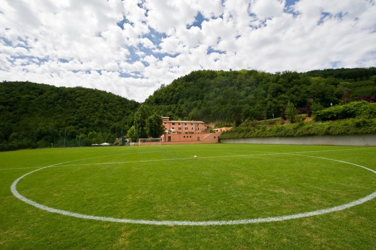 Campo da calcio circondato da colline verdi e uno stabile in lontananza.