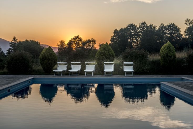 Un tramonto tranquillo si riflette nella piscina, contornata da sdraio e piante verdi.