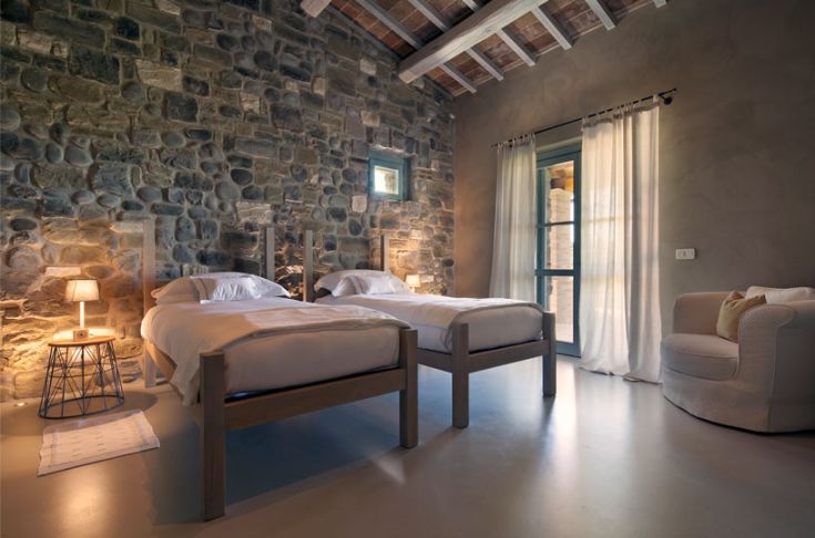 Chambre lumineuse avec des murs en pierre, des lits individuels et une fenêtre donnant sur un espace vert.