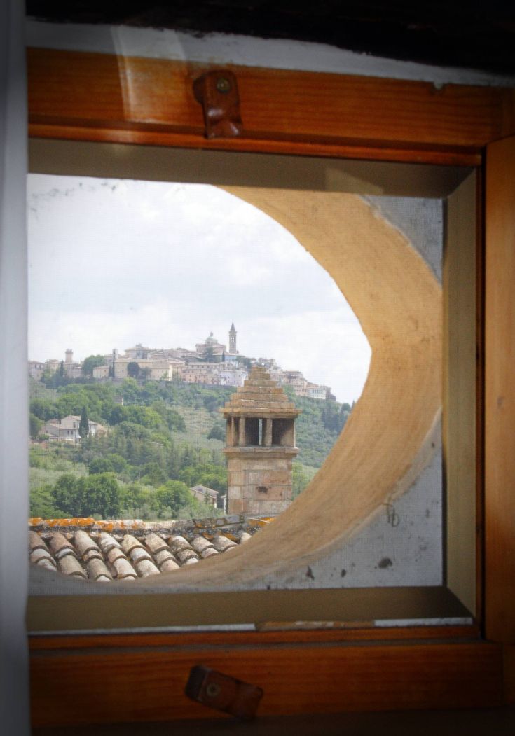 Scene of a hilly village seen through a circular window, featuring buildings and trees typical of the Umbrian landscape.