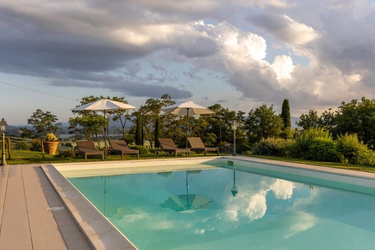 A pool nestled in the greenery of a rural Umbrian area, surrounded by loungers and umbrellas, creating a tranquil spot.