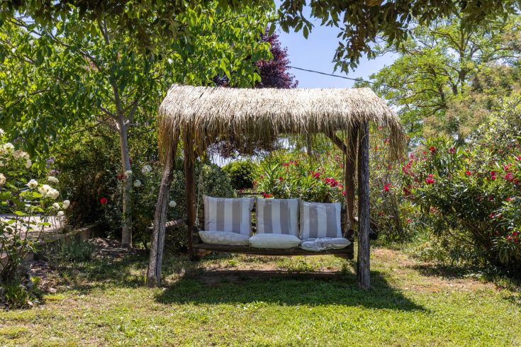 A simple gazebo with cushions, surrounded by a green garden and colorful flowers.