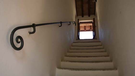 Scala bianca con corrimano in ferro che porta a una porta aperta, illuminata dalla luce del giorno.