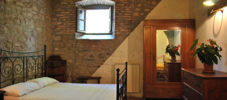 Camera semplice con pareti in pietra e letto matrimoniale; finestra aperta su un ambiente sereno.