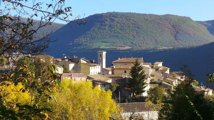 Scena di un borgo circondato da verde, con edifici storici e colline sullo sfondo.