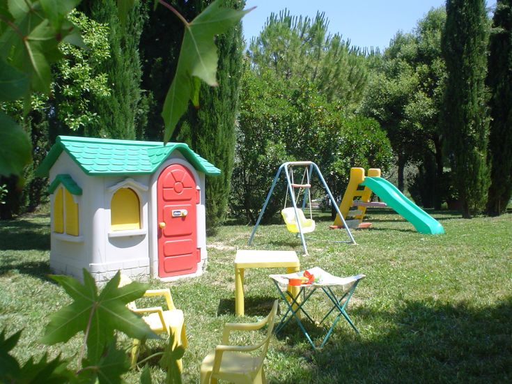 Un terrain de jeux avec un toboggan, une balançoire et une petite maison, entouré d'arbres et d'herbe verte.