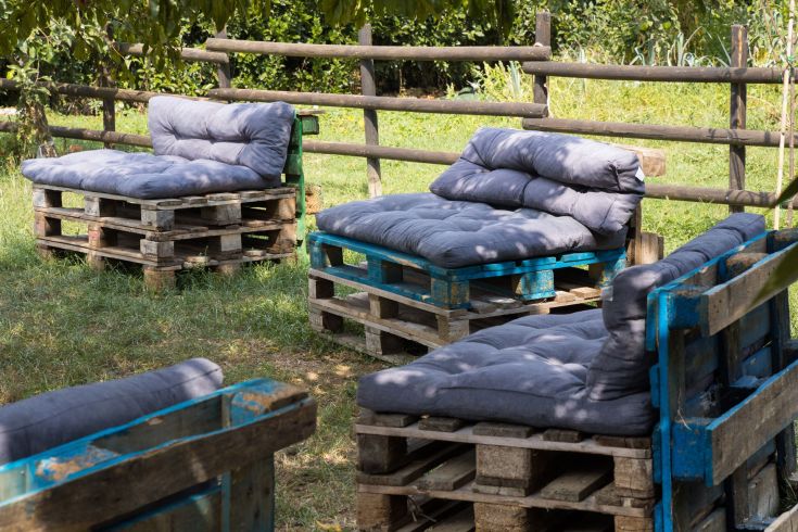 Divani in legno costruiti con pallet e dotati di cuscini, collocati in un ambiente verde e naturale.