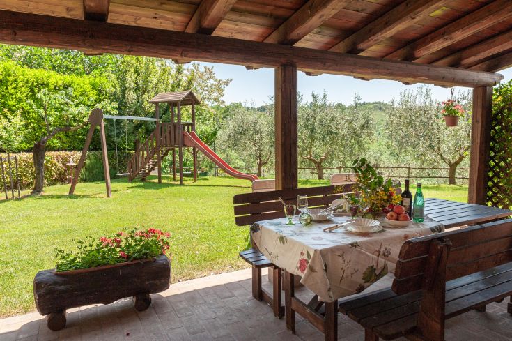 Spazio esterno attrezzato con tavolo, circondato da ulivi e un'area per bambini.