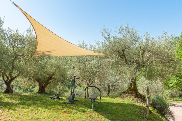 Un'area fitness all'aperto ombreggiata da un tendone, immersa in un contesto naturale con ulivi.