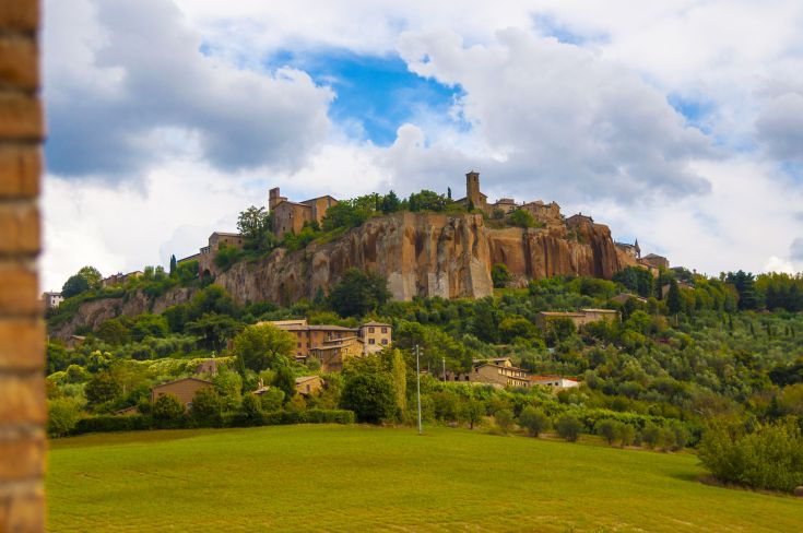 Scena di un antico borgo in collina, immerso nel verde e con nuvoloso cielo.
