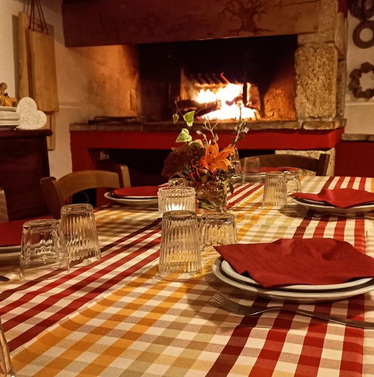 Table bien mise avec nappe à carreaux, verres et décorations florales, près d'une cheminée allumée.