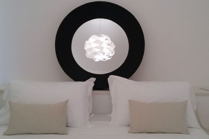 Ein einfaches Bett mit schwarzem Kopfteil und einer weißen Lampe darüber, in einem minimalistischen Raum.