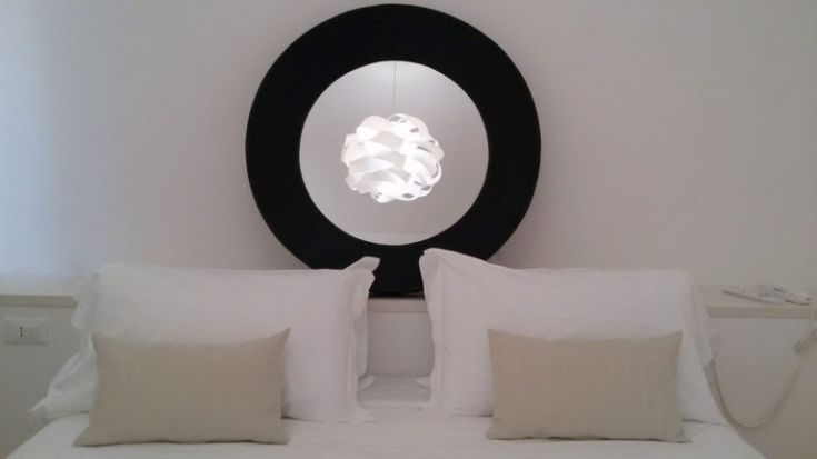 Einfaches Schlafzimmer mit einem weißen Bett, hellen Kissen und einer zeitgenössisch gestalteten Lampe.