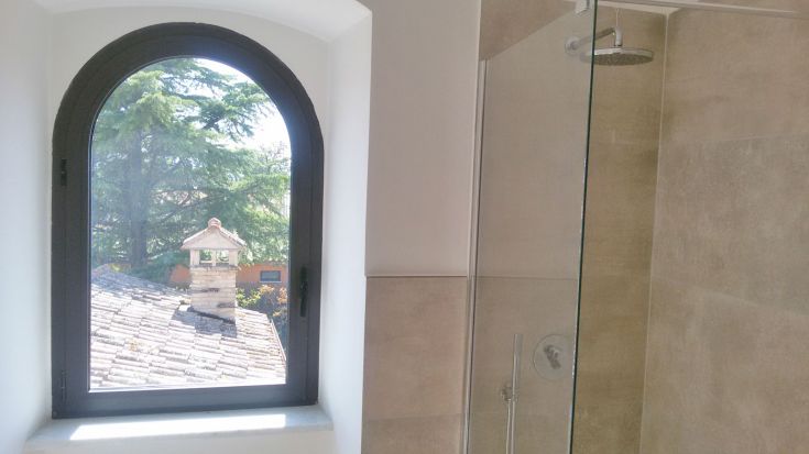 Geräumiges Badezimmer mit Dusche, ausgestattet mit einem Fenster, das auf Bäume und umliegende Dächer blickt.