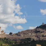 Historische Stadtansicht mit alten Gebäuden und grünen Hügeln im Hintergrund.