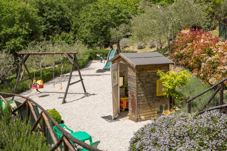An outdoor playground featuring swings, a slide, and a playhouse, surrounded by green space.