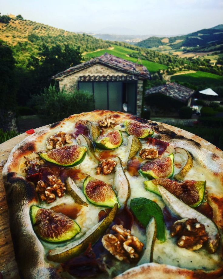 A pizza topped with figs, pears, and walnuts, set in a picturesque Umbrian countryside.