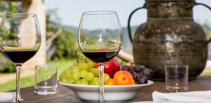 Two glasses of red wine next to fresh fruit and an old pitcher, set in a natural environment.