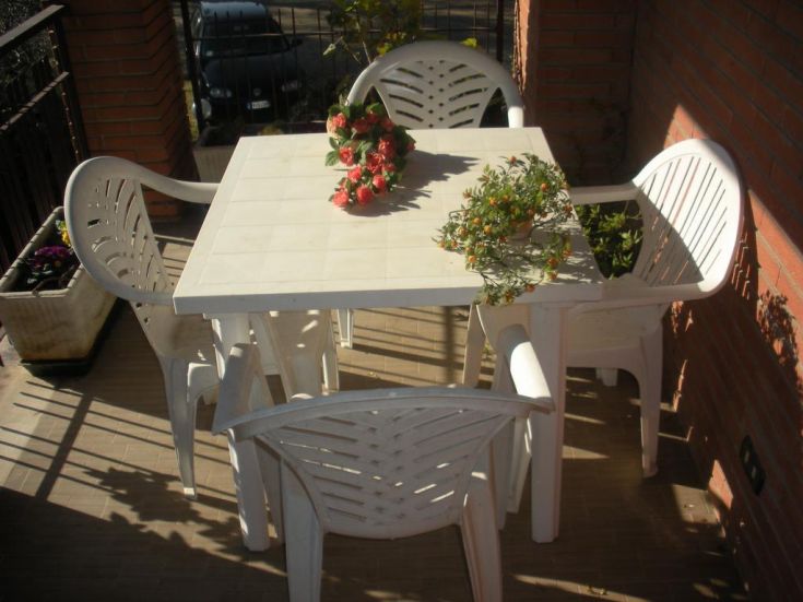 A sunny terrace with a white table and chairs, decorated with green plants and colorful flowers.