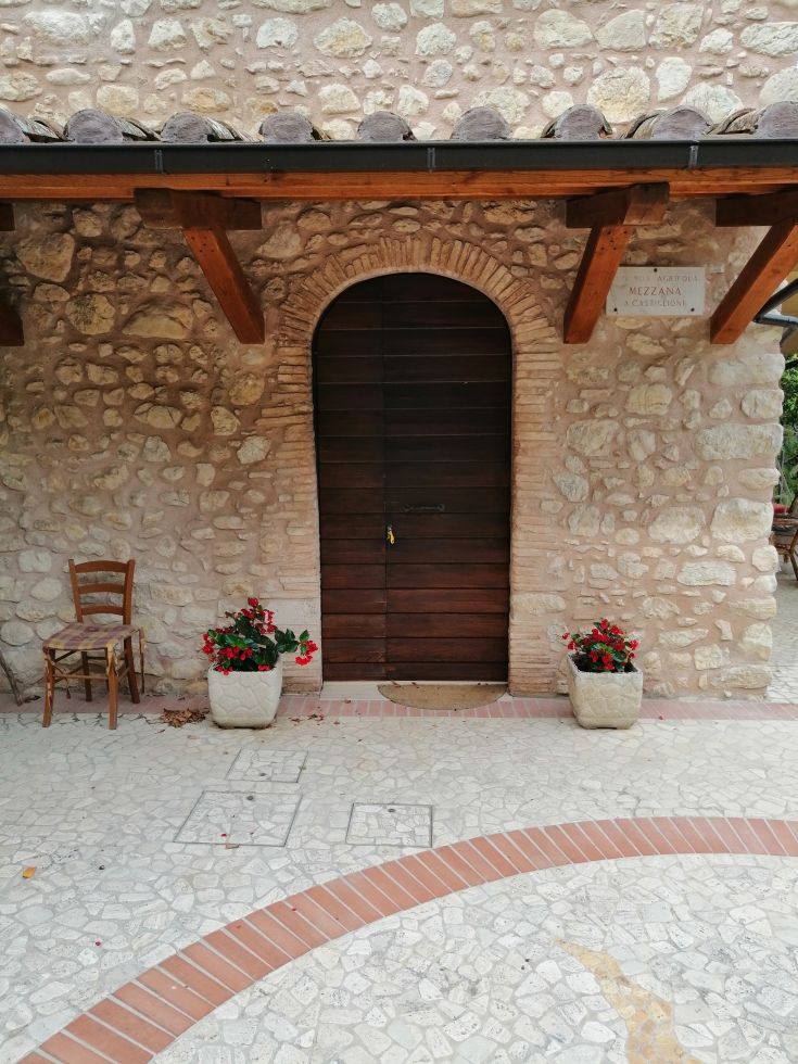 A renovated farmhouse with a garden, situated in the green nature of Valnerina. Features a wooden door and flowering plants.