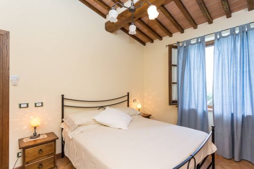 Bright matrimonial room with a large bed, soft lighting, and a window adorned with blue curtains.