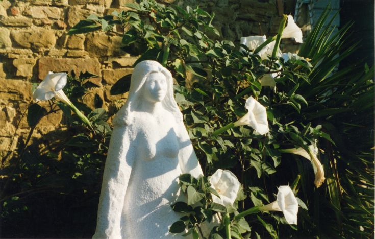 Scultura bianca collocata tra piante e fiori bianchi in un contesto naturale.