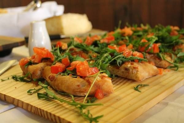 Focaccia préparée avec des tomates fraîches et de la roquette, présentée sur une planche en bois.
