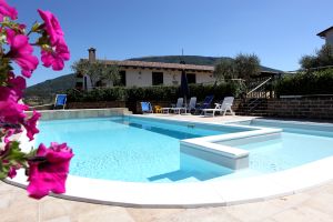 Una piscina circondata da un giardino con piante e fiori, ideale per momenti di relax. Sullo sfondo si intravede un edificio rustico.
