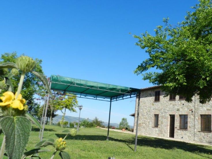 Szene eines Bauernhauses in Umbrien mit einem gut gepflegten Garten und einer Laube unter einem klaren Himmel.