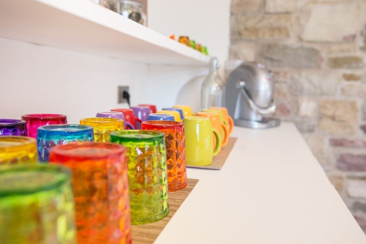 Bicchieri e tazze di diversi colori organizzati su uno scaffale, offrono una nota vivace all'ambiente della cucina.