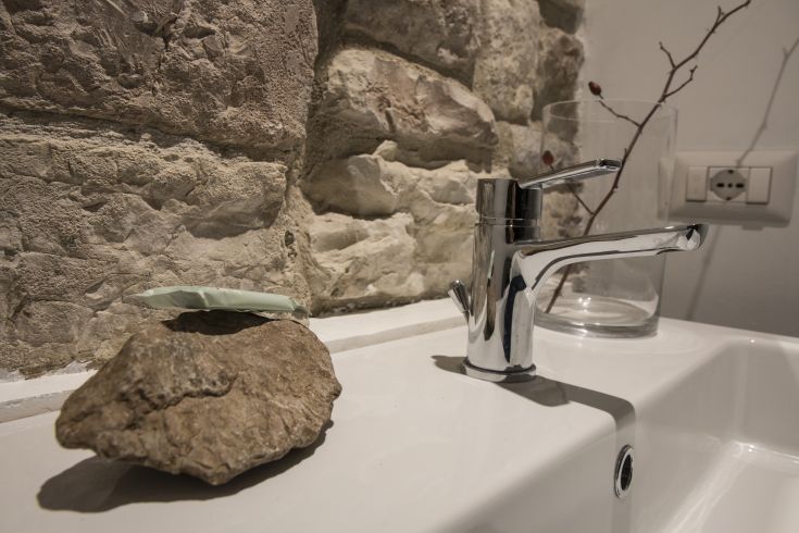 A sleek sink with a shiny faucet, beside a stone rock and green soap, in front of a stone wall.